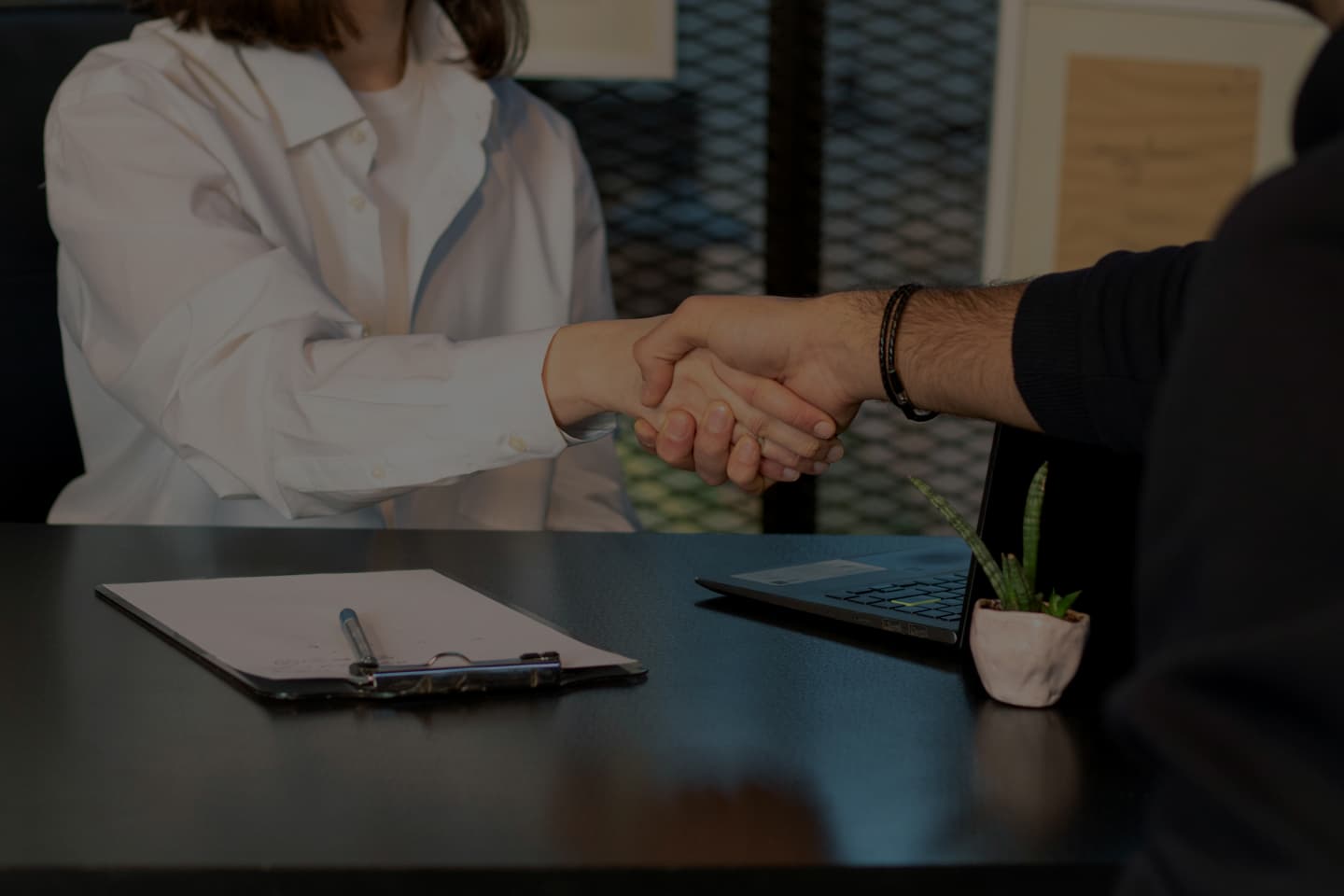 Two people shaking hands across a desk, dimmed